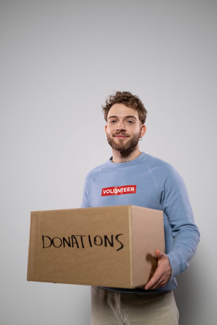 gallery-1 Smiling young man in a volunteer shirt holding a cardboard box labeled donations.
