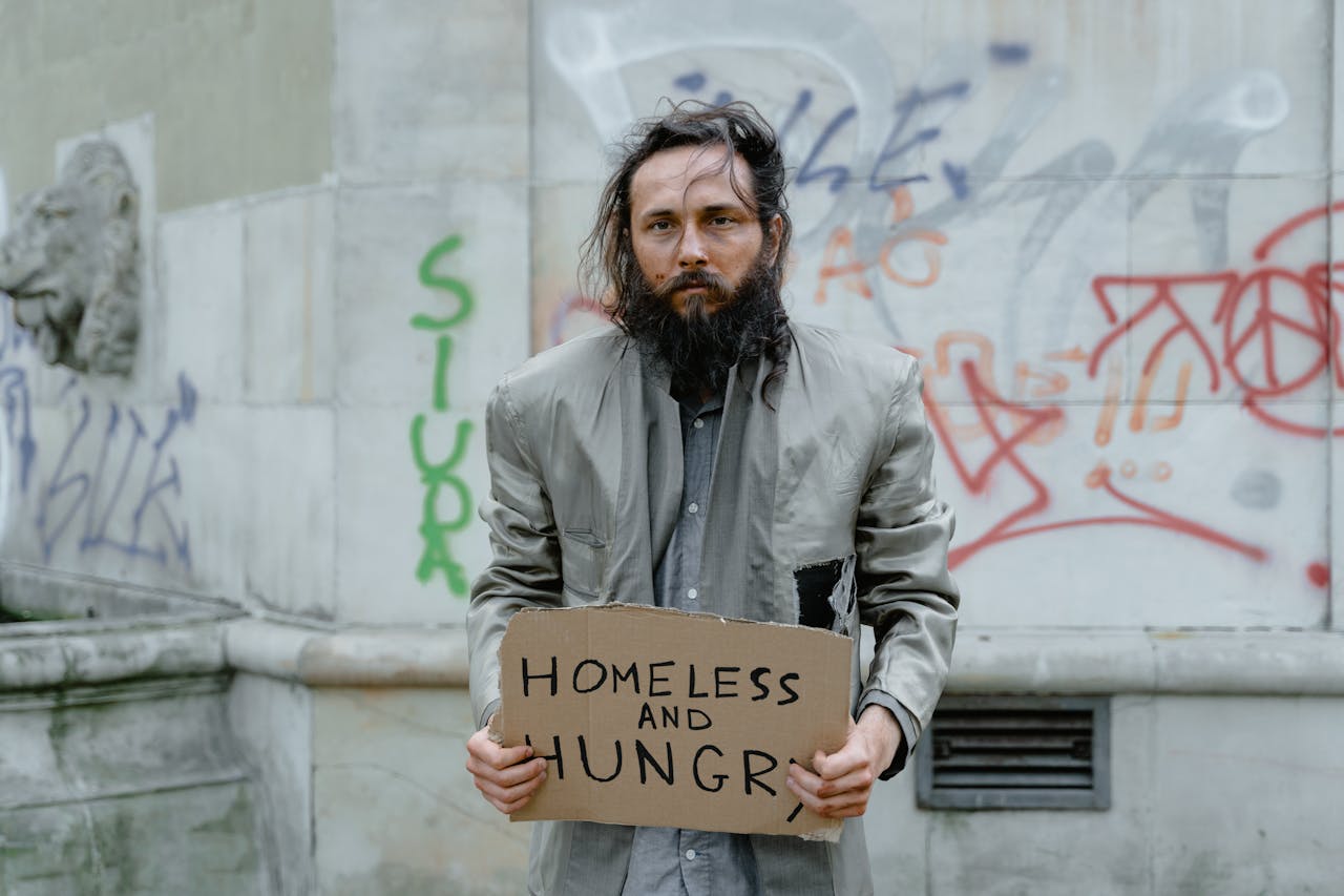Homeless man standing on the street holding a sign, with graffiti in the background.